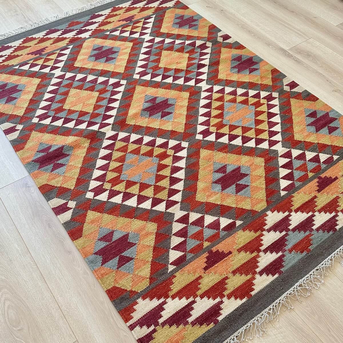 Covor subtire cu tesatura plata Kilim Alwar cu motive traditionale in culori calde de rosu maro si portocaliu in doua dimensiuni, model geometric in romb, pe podea